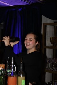 A young woman smiles while shaking a cocktail shaker behind a bar.