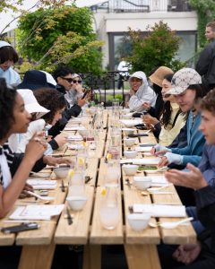 A long wooden table is set with bowls and glasses, filled with a diverse group of people enjoying a meal outdoors.