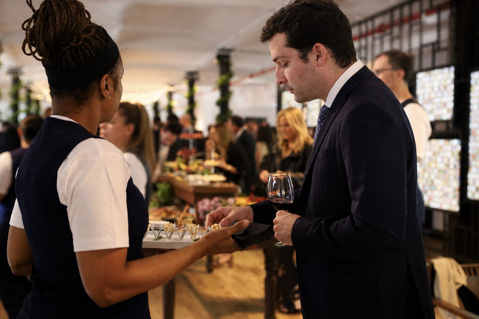 A man in a suit examines a platter of appetizers being offered by a server at a lively event.