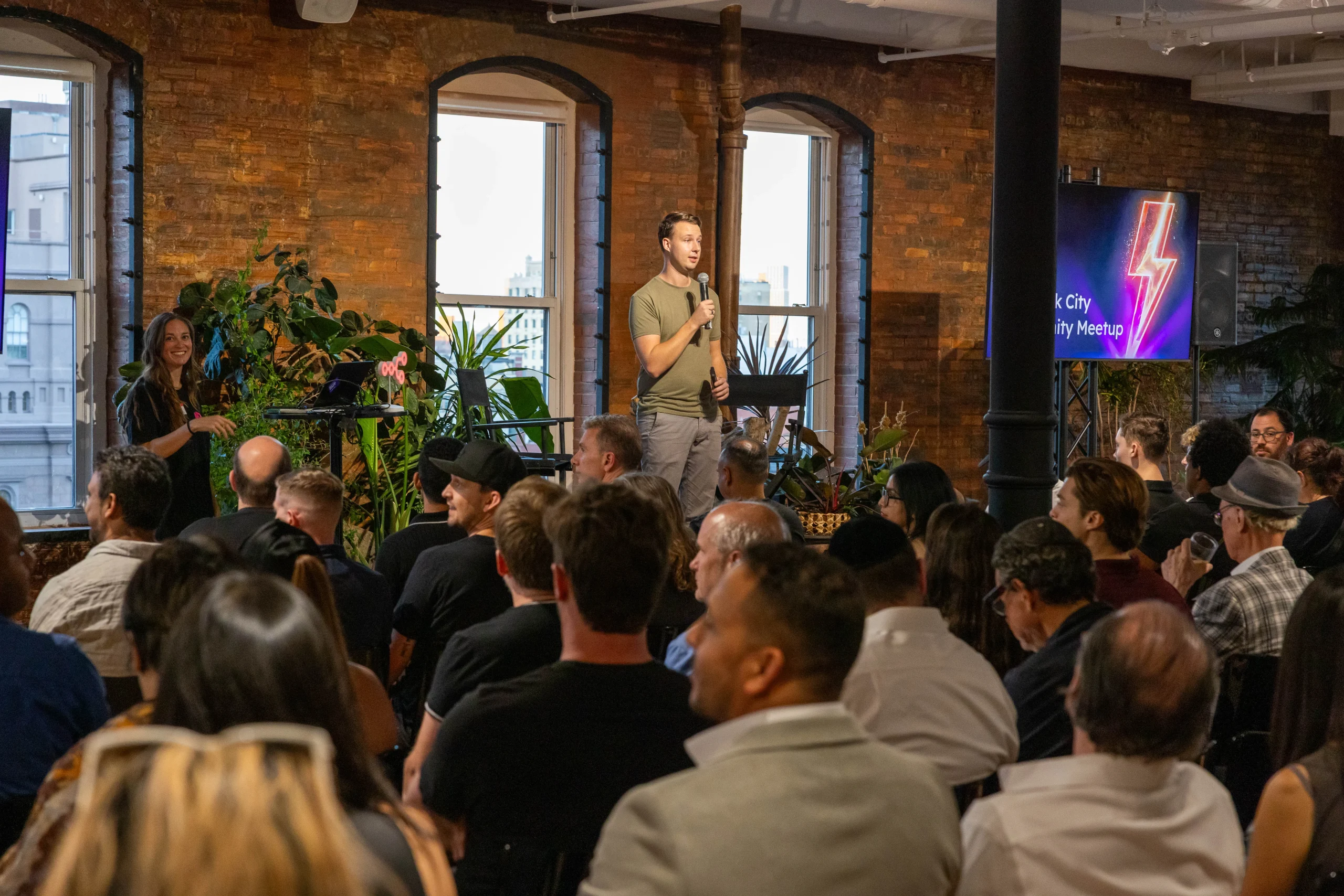 A speaker addresses an audience during a meetup event in a stylish venue with large windows and plants.
