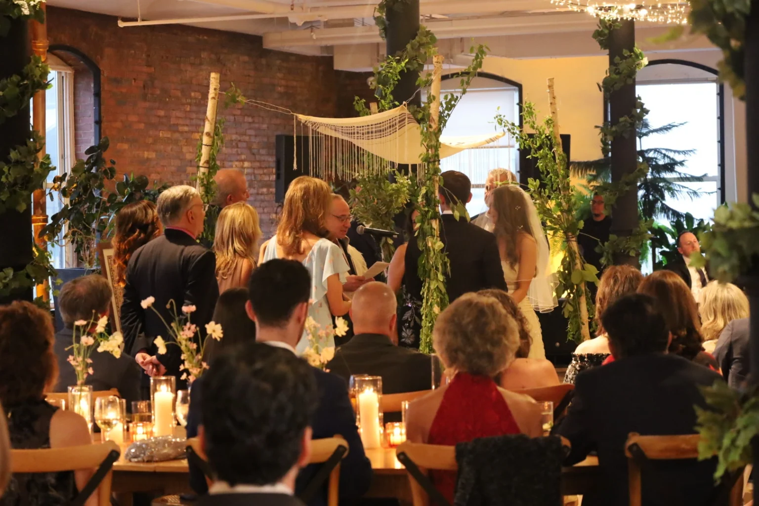 A wedding ceremony taking place indoors with guests seated and an officiant addressing the couple under a floral canopy.