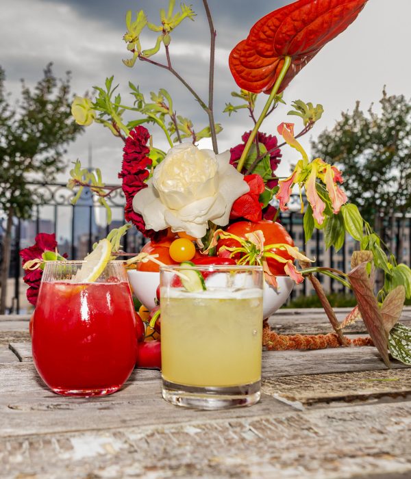 Two colorful cocktails are placed on a rustic wooden table beside a vibrant floral arrangement against a cloudy sky.