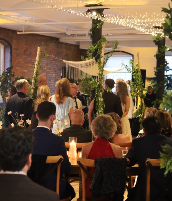 A wedding ceremony taking place indoors, surrounded by guests seated at a long table with candles and flowers.