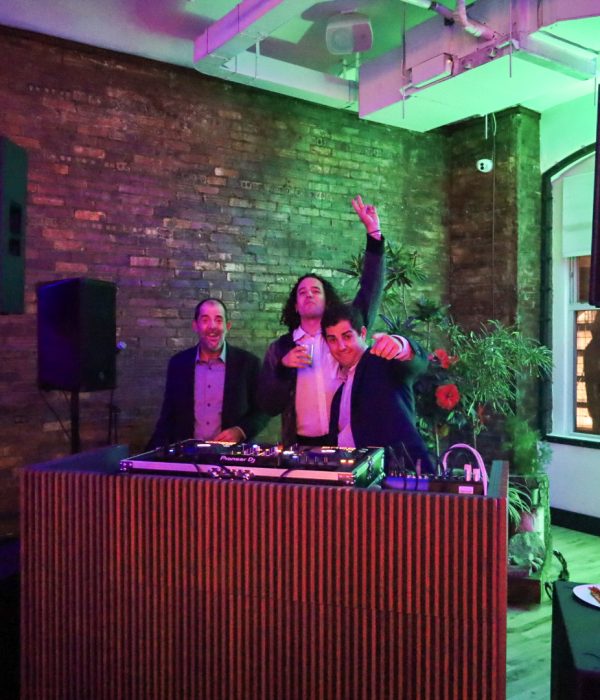 Three men pose joyfully at a DJ booth in a vibrant, colorful setting.